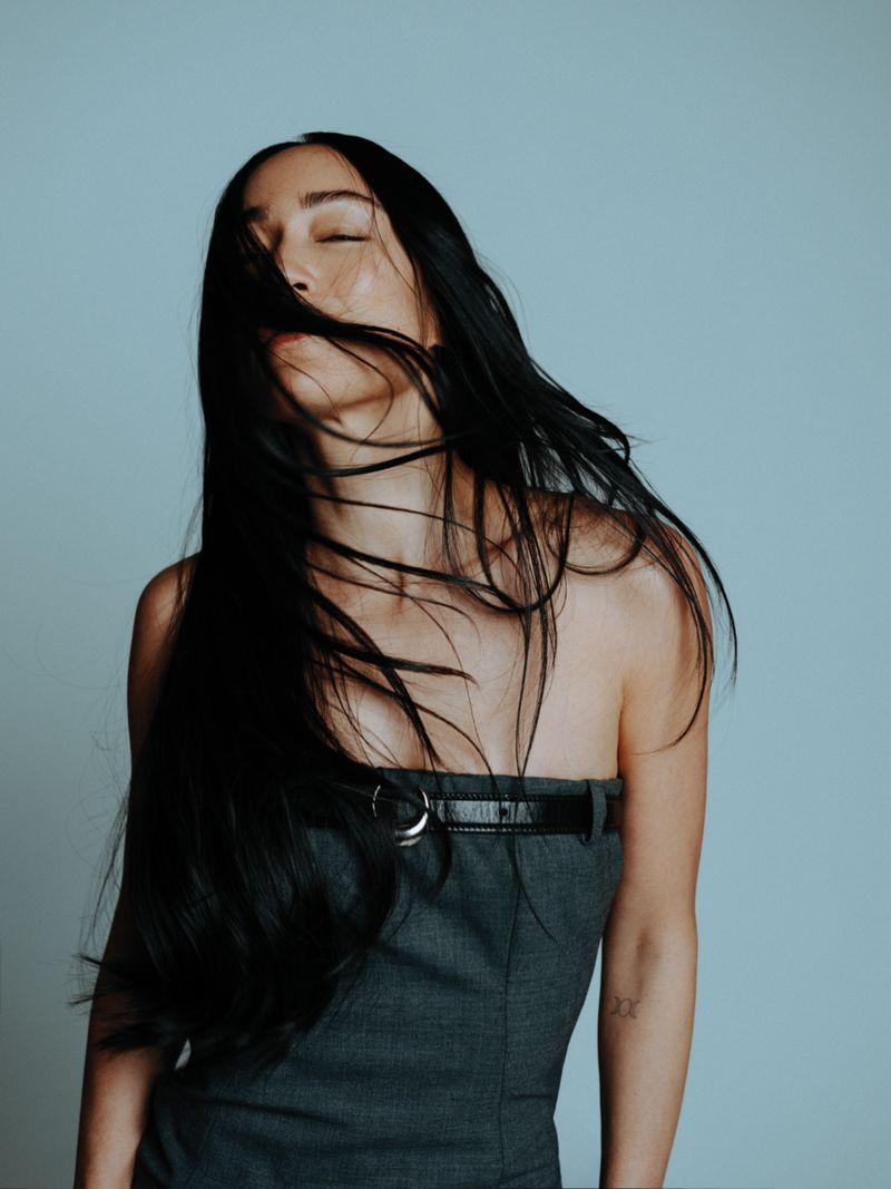 Portrait of a woman with long black hair blowing in the wind, wearing a strapless grey dress with a black belt, against a light blue backdrop.