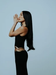 Profile portrait of a woman with long black hair wearing a black outfit with gold trim, posing with hands together