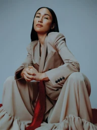 Woman wearing a beige blazer and pleated trousers squatting against a grey studio backdrop for Schön Magazine.