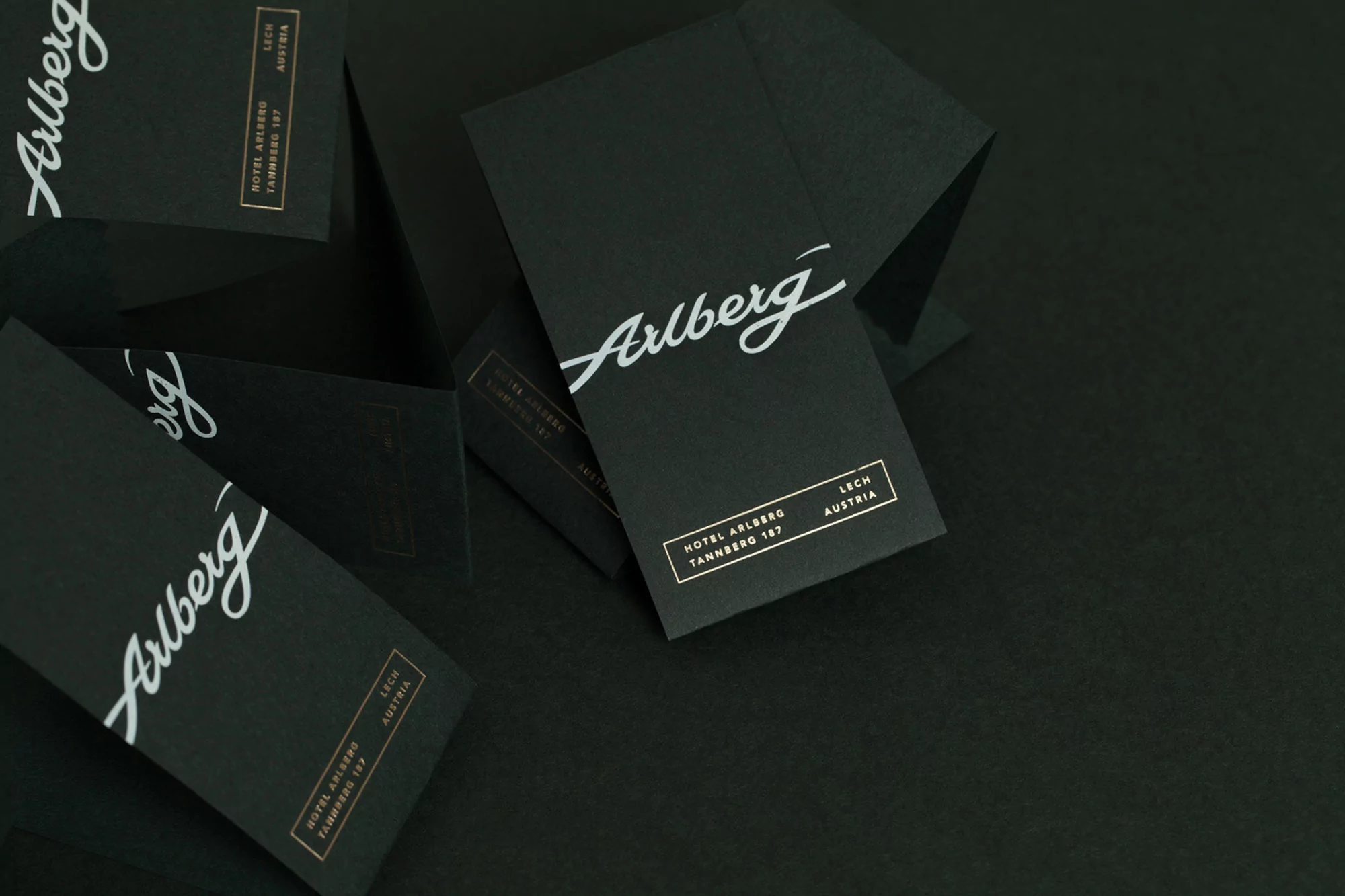 A pile of dark business cards with white cursive text "Arlberg" and smaller bronze text listing hotel and location details.