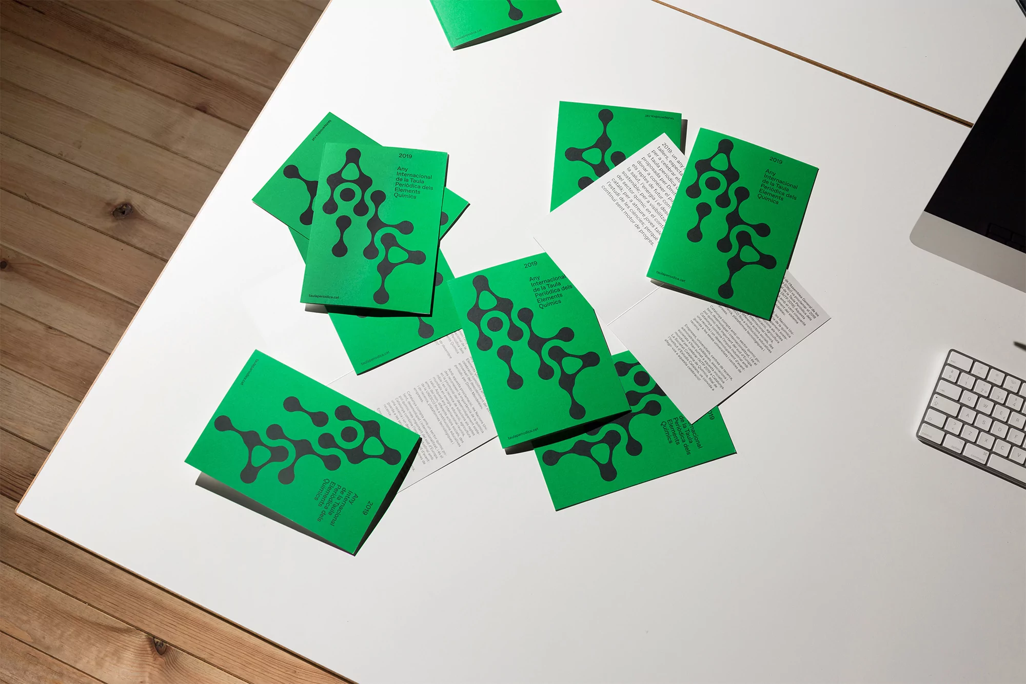 Overhead shot of green brochures scattered on a white table with a computer and keyboard, minimal design, and bright lighting.