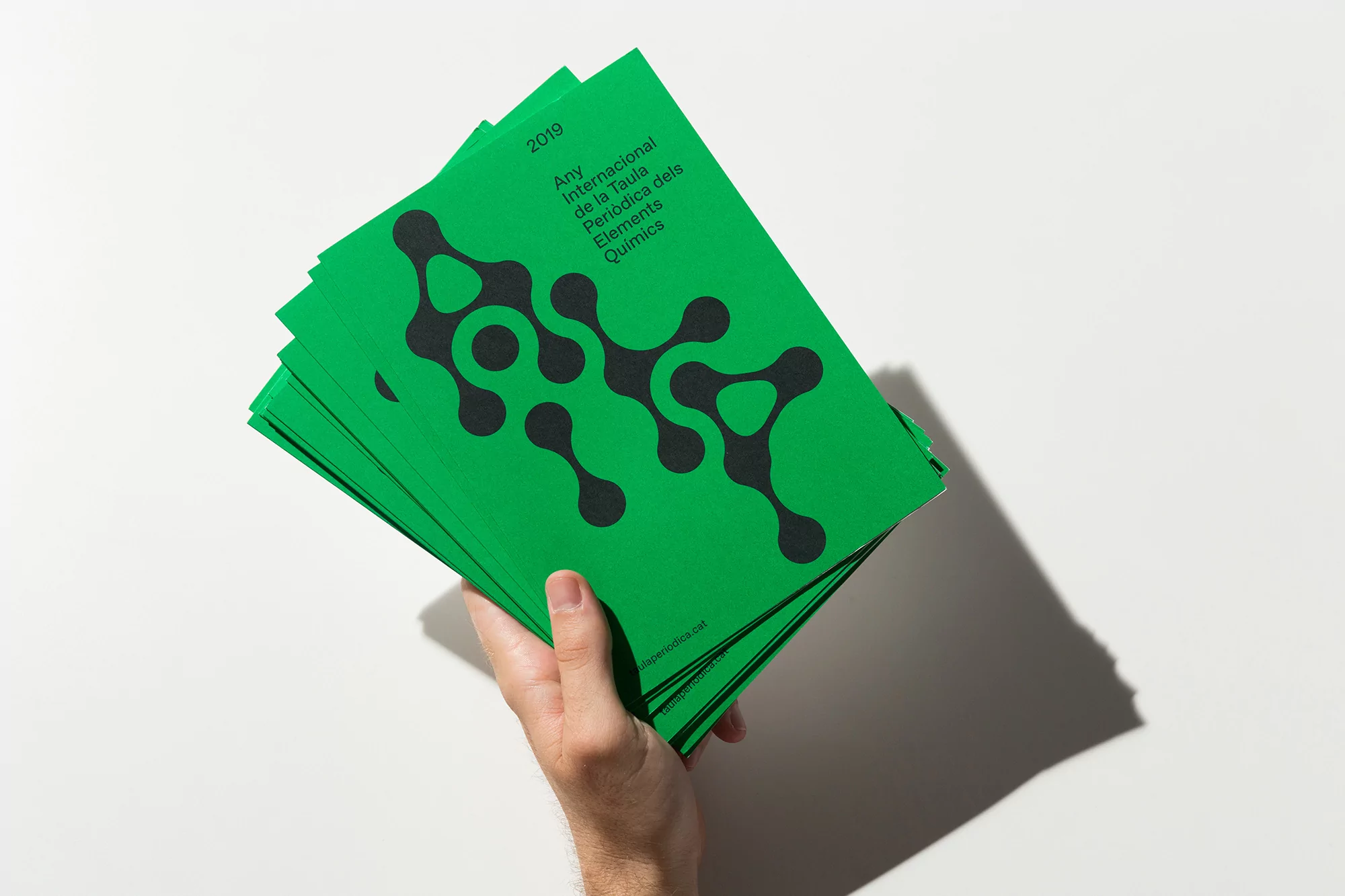 Hand holding a stack of green booklets with a black graphic design, promoting the International Year of the Periodic Table.