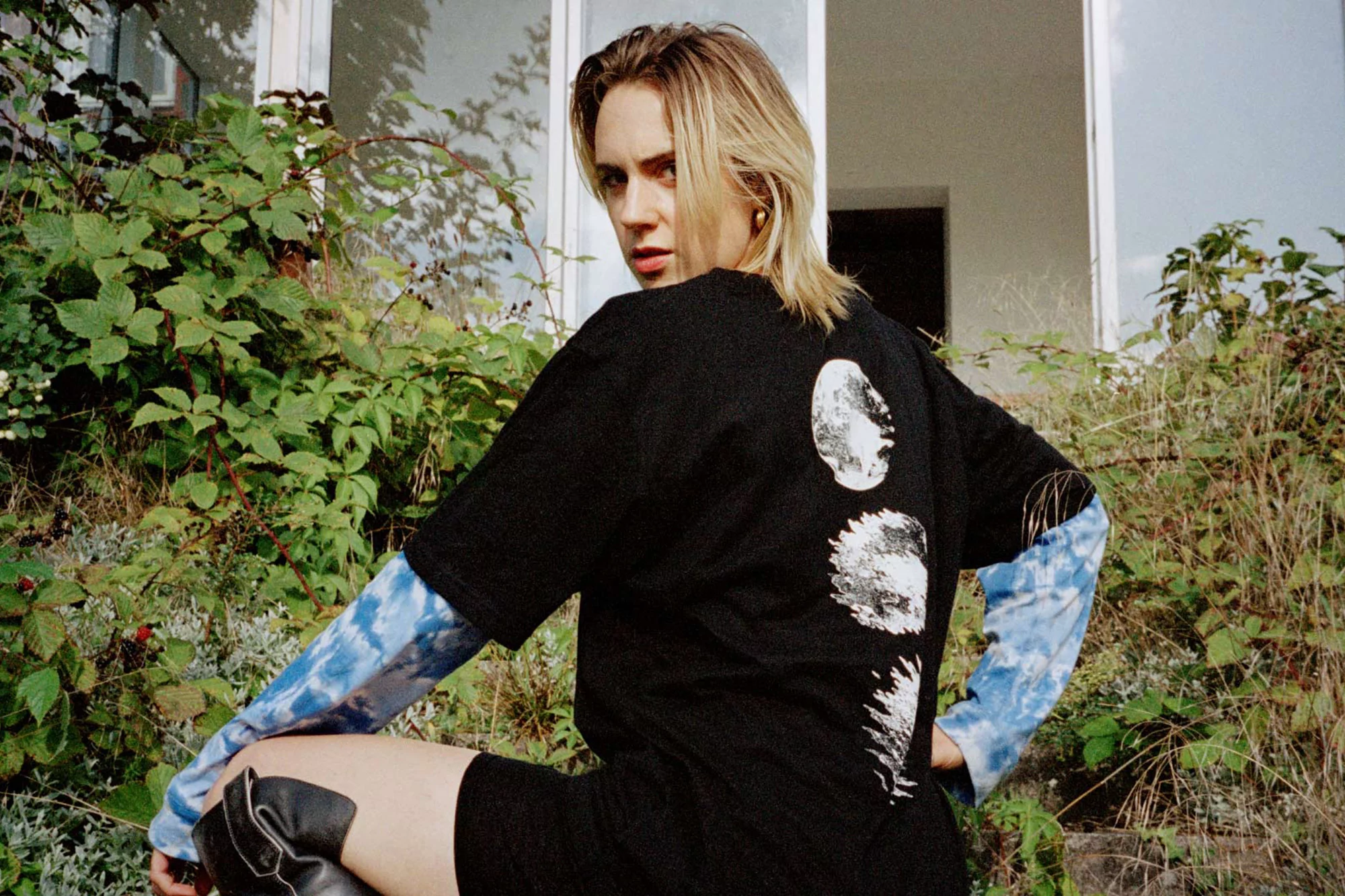 Young woman poses in black t-shirt with moon phases graphic, black shorts and boots, outdoors with green foliage in the background.