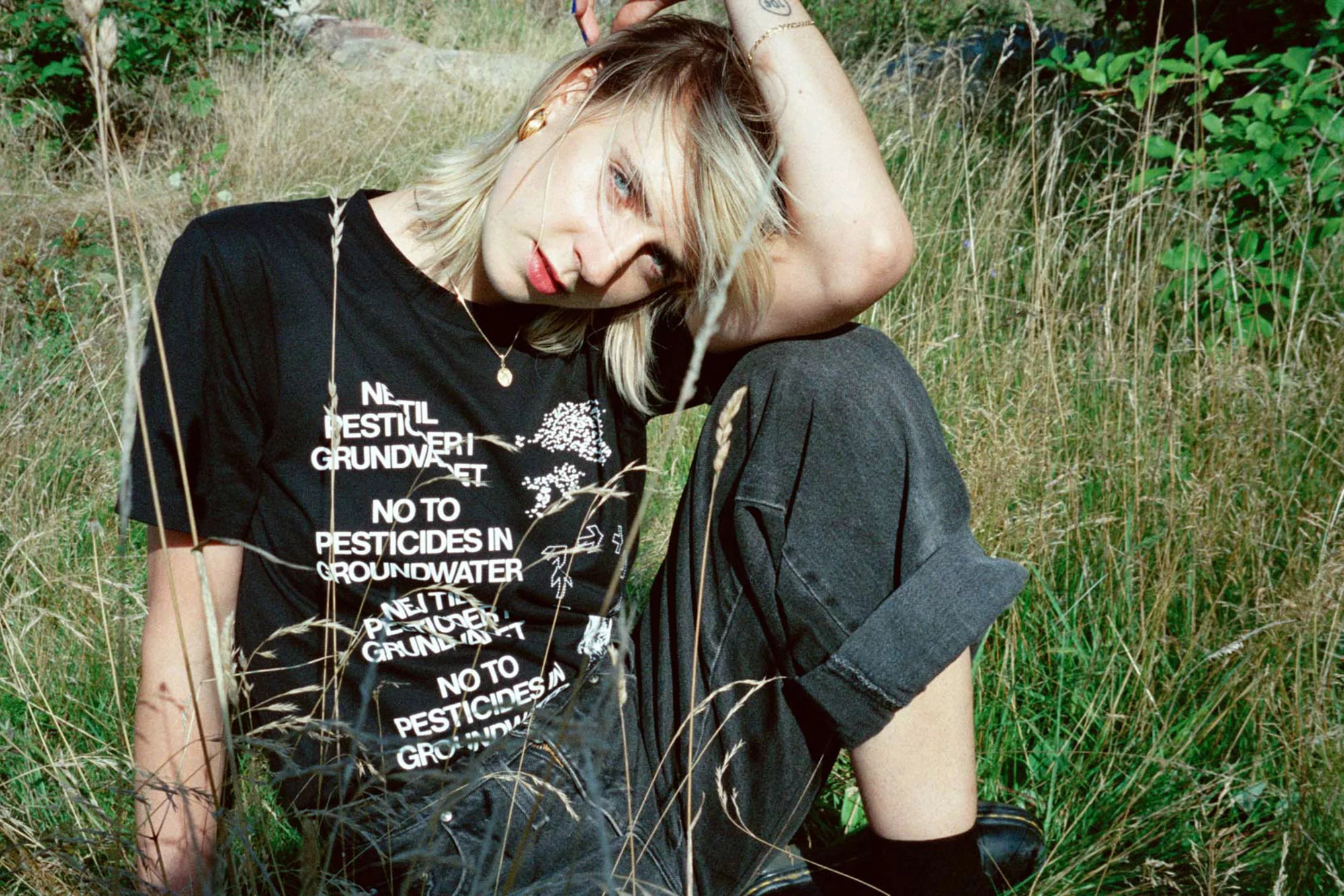 Young adult woman with blonde hair sits in a grassy field, wearing a graphic tee and jeans, looking at the camera in an outdoor setting.