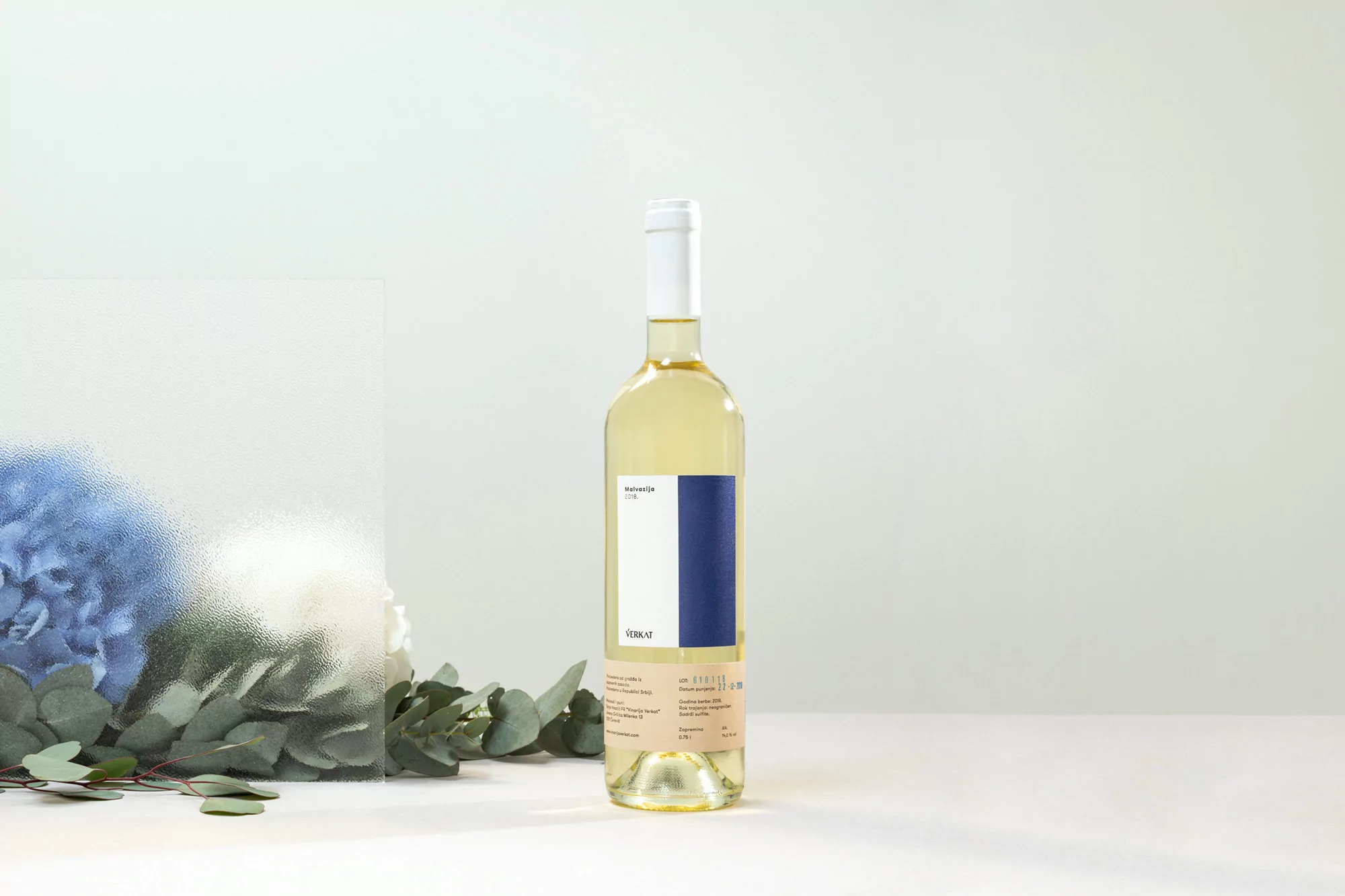 Wine bottle with white wine, featuring a white label with blue stripe, set against a clean background with flowers and leaves.