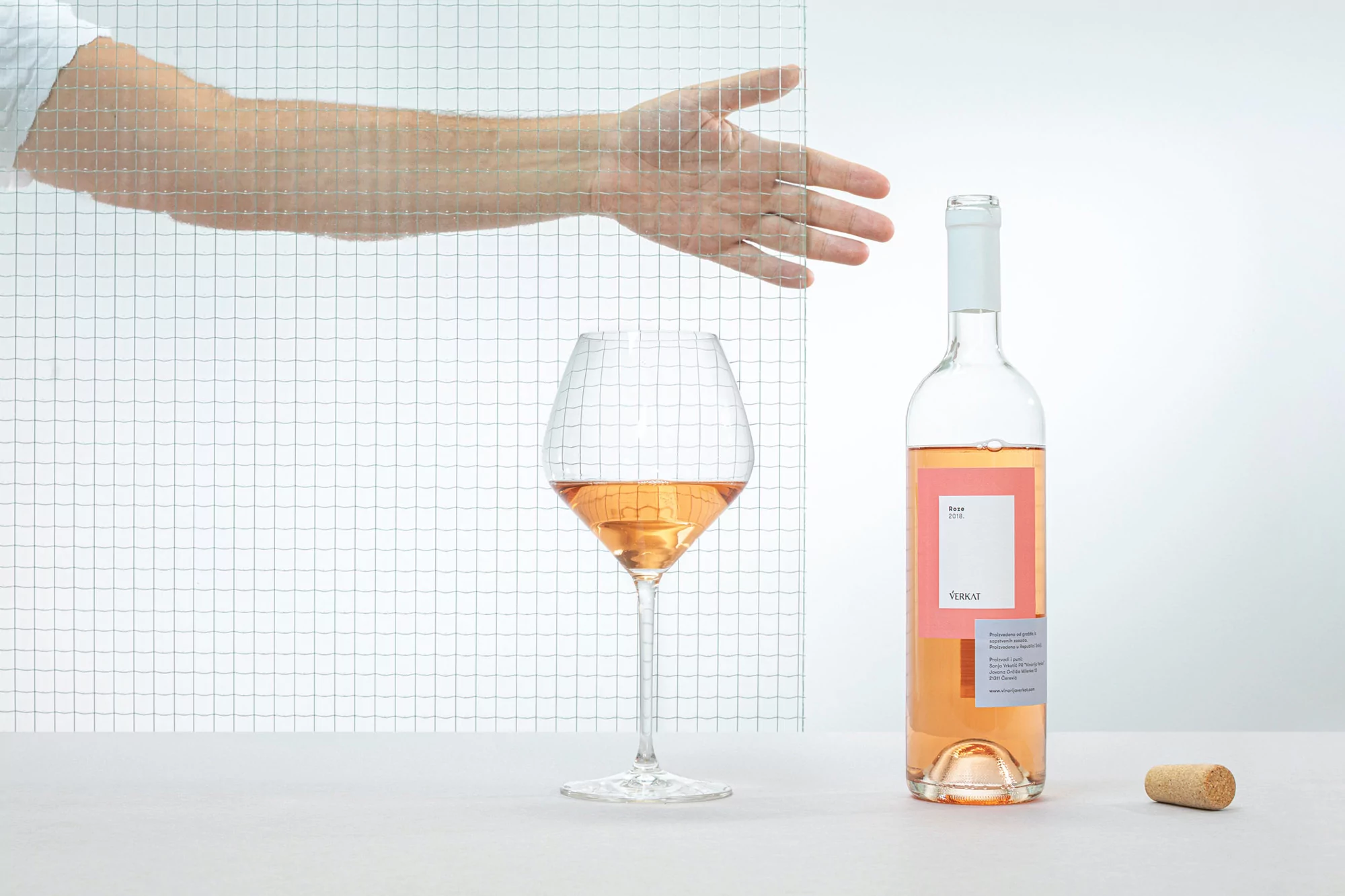 Still life of rosé wine bottle and glass, against a white background with a grid-patterned panel and a human hand reaching in.