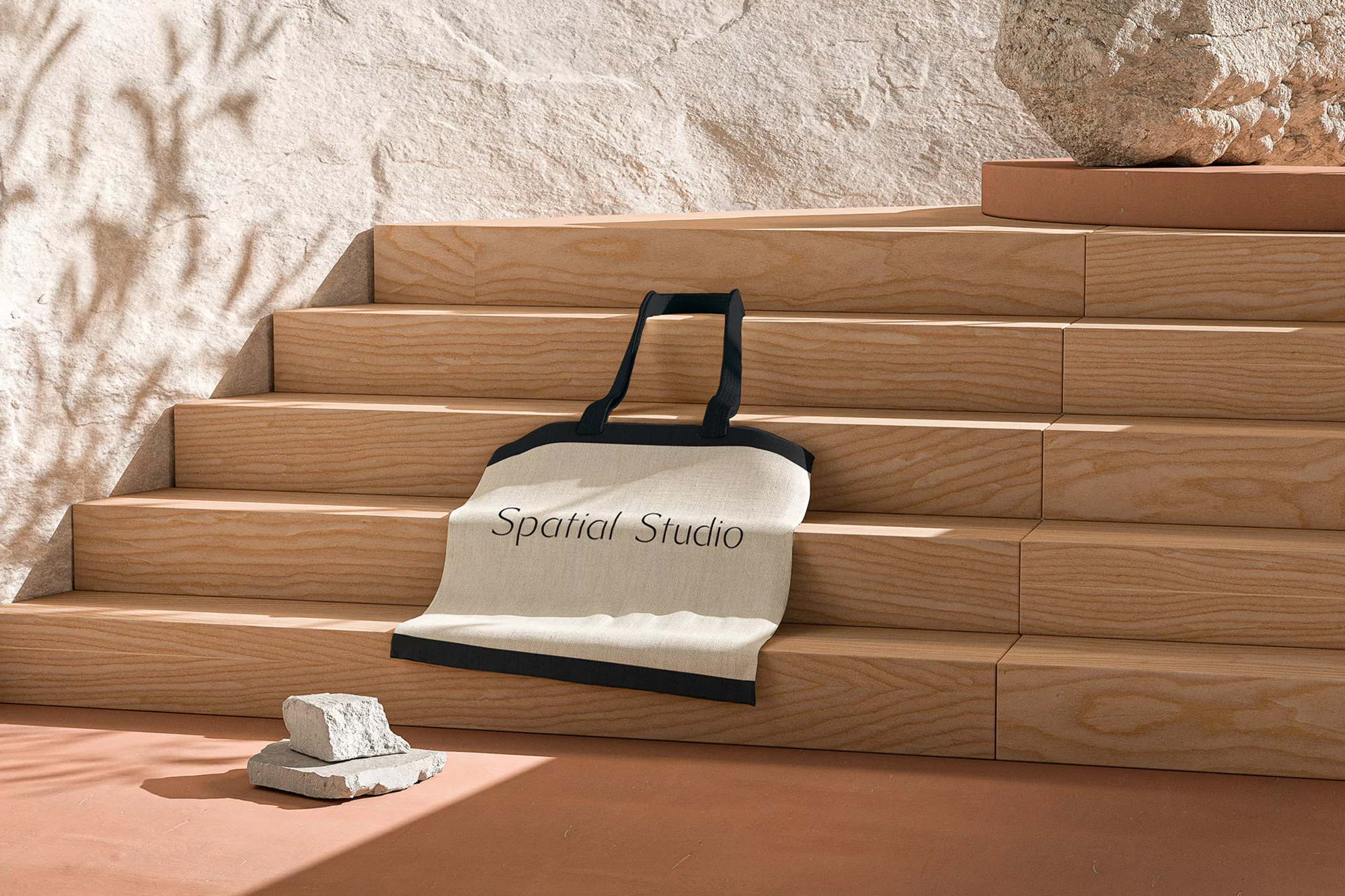 Beige tote bag with black trim on wooden steps near stone objects in a minimalist studio setup with warm lighting and natural textures.