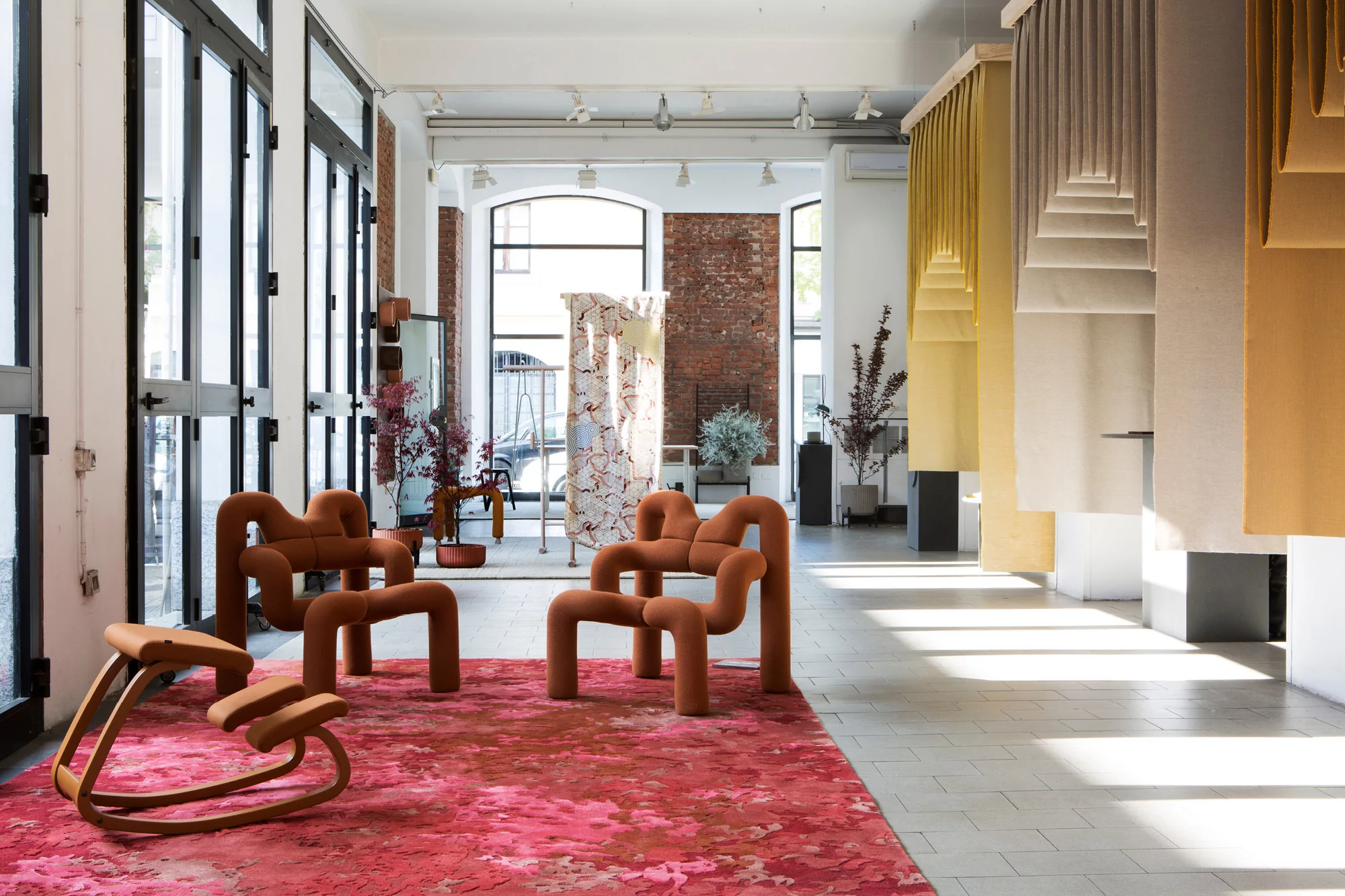 Interior design showroom featuring uniquely shaped chairs and stools displayed on a red patterned rug. Natural light fills the space.