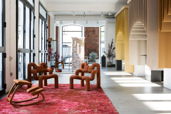 Interior design showroom featuring uniquely shaped chairs and stools displayed on a red patterned rug. Natural light fills the space.