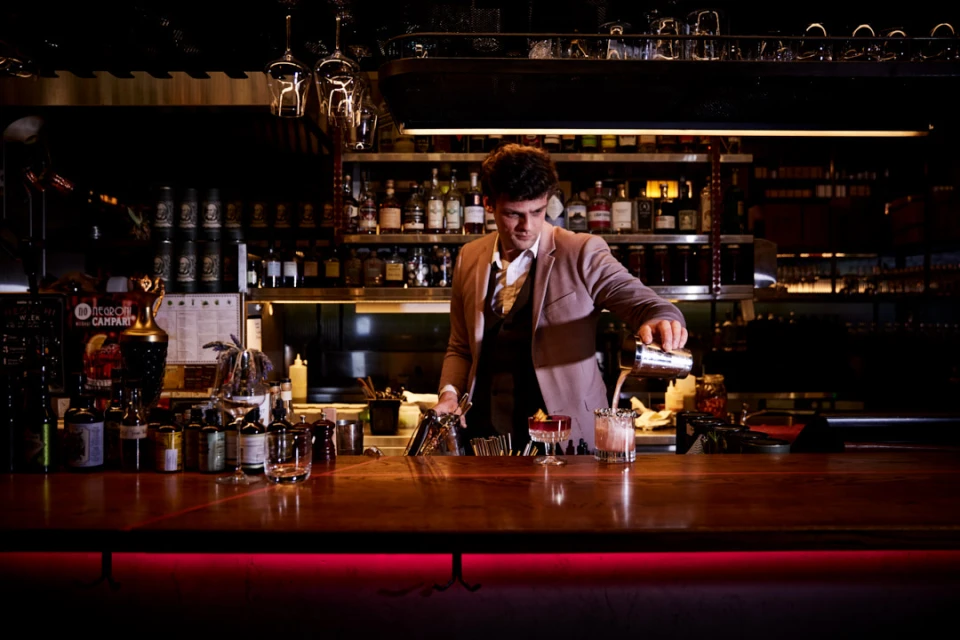 Bartender in a suit mixing a cocktail at a bar with an array of bottles on shelves in the background, illuminated by warm lighting.