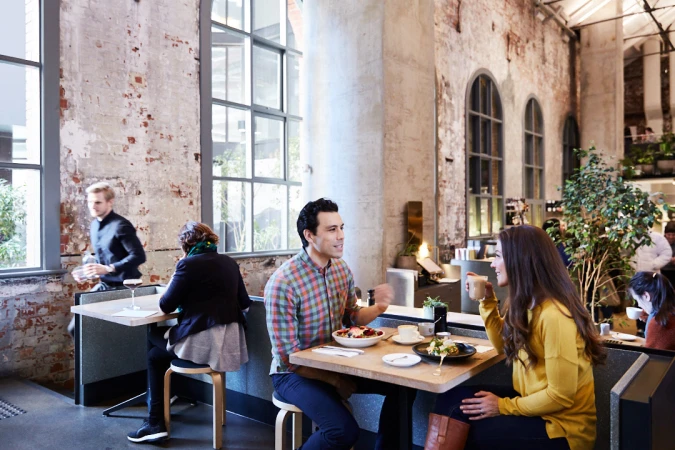 People dining in a restaurant with exposed brick walls and large windows, enjoying meals at wooden tables with modern seating.