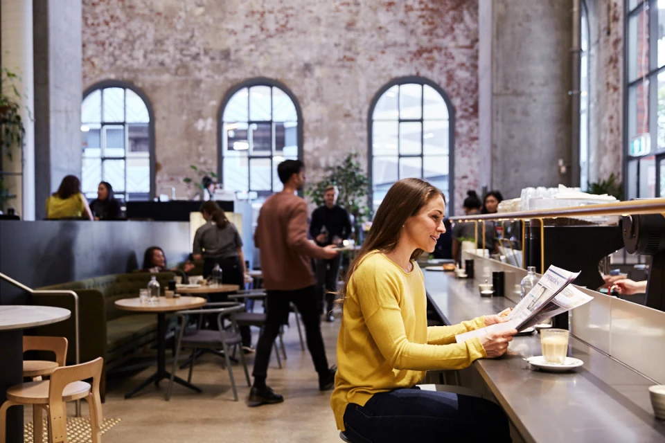 Woman in yellow sweater reads newspaper at coffee shop counter while people sit at tables in the background of industrial-style interior.