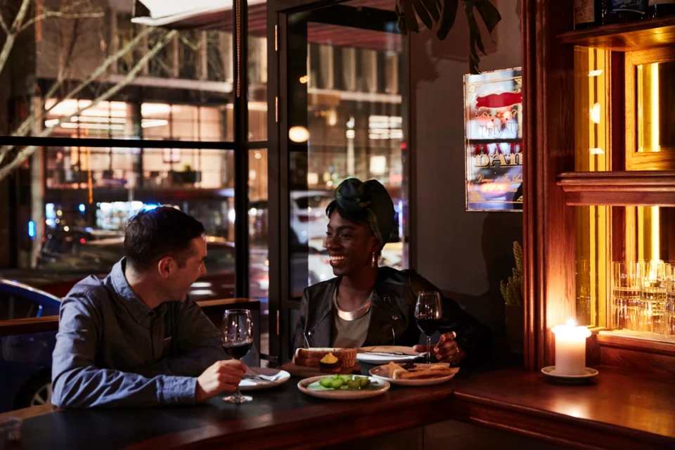 A couple sits at a cozy restaurant table at night, sharing wine and appetizers in warm lighting with city lights visible outside.
