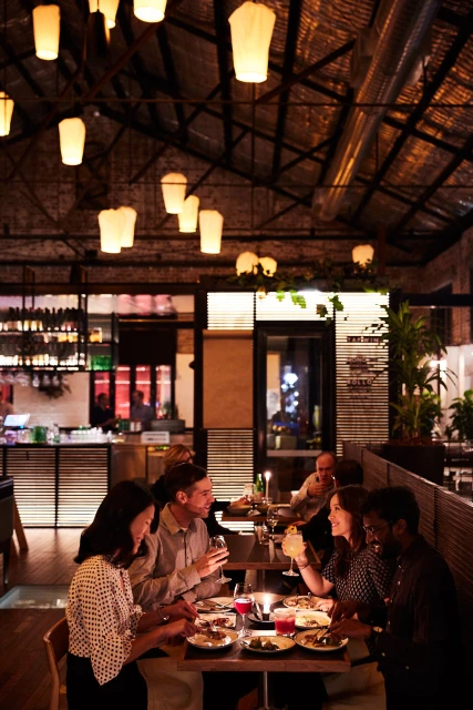 People dine at a warmly lit restaurant with paper lantern lighting, sharing food and drinks at their tables in an intimate setting.
