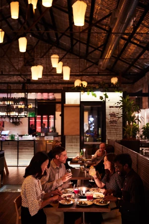 People dine at a warmly lit restaurant with paper lantern lighting, sharing food and drinks at their tables in an intimate setting.