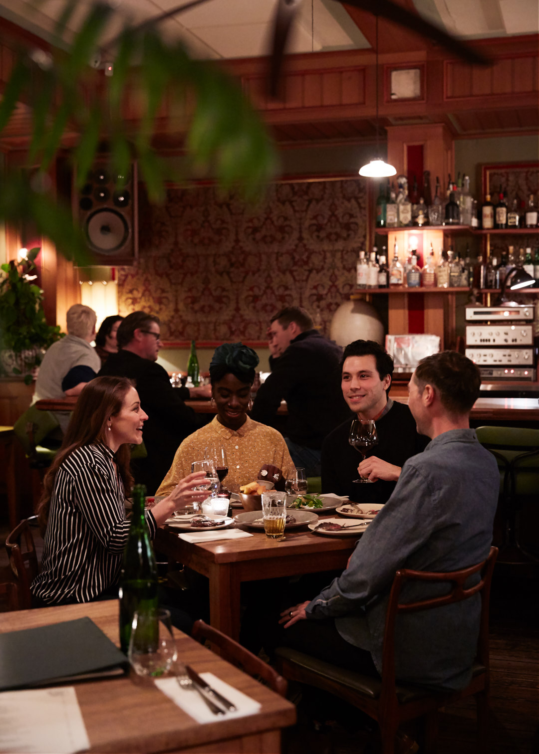 Group of four adults sit at a table in a restaurant, enjoying a meal with drinks, soft lighting, and a backdrop of other restaurant patrons.