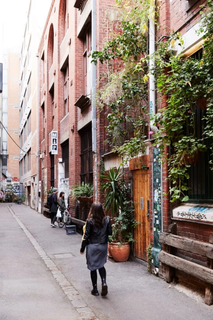 Woman walks down a narrow urban alleyway with brick buildings and green plants, creating a vibrant city scene.