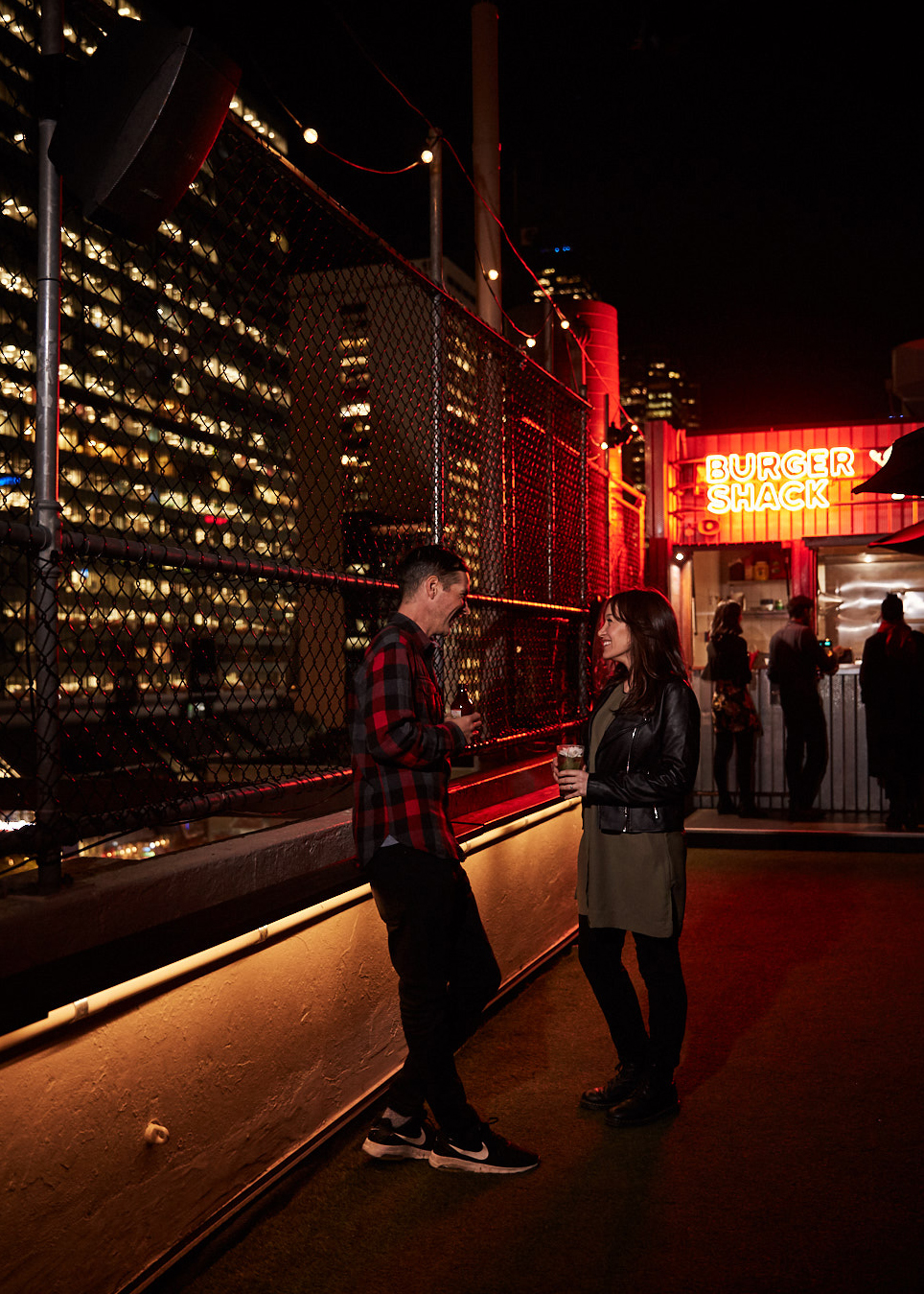 A man and woman in a nighttime urban setting, possibly a rooftop, conversing near a food stand with city buildings in the backdrop.