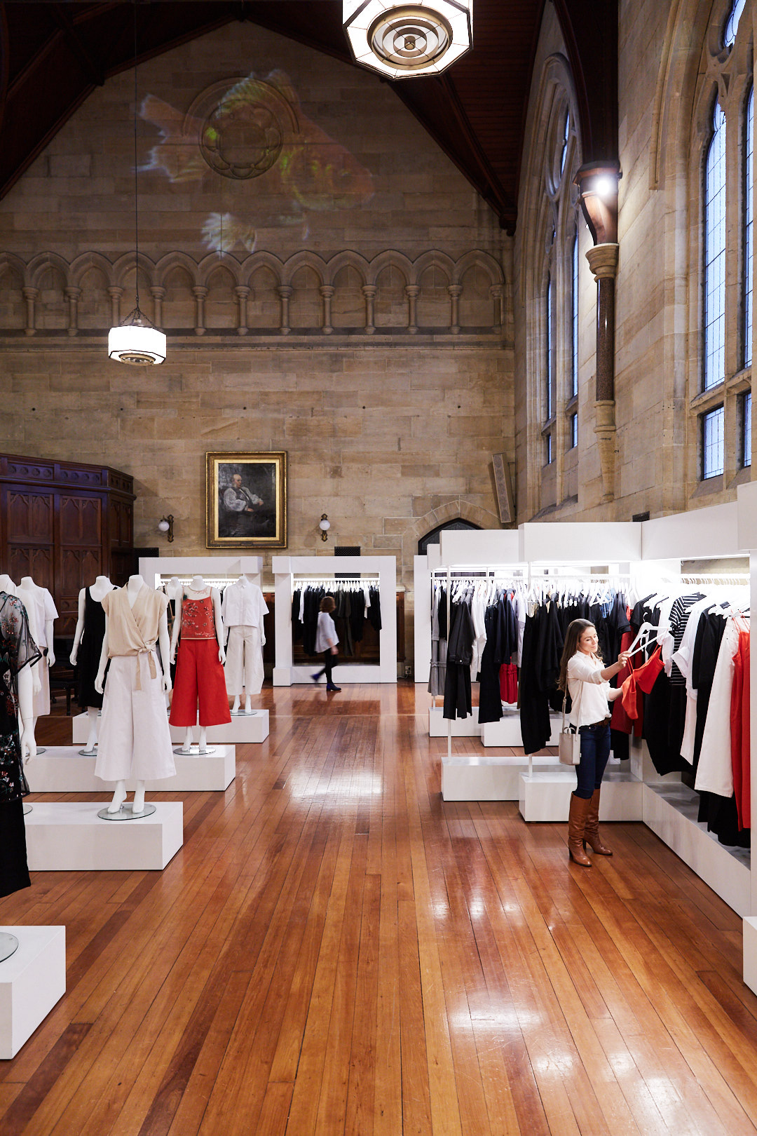 Clothing on mannequins and racks is displayed in a showroom with hardwood floors, arched windows, and a high ceiling.