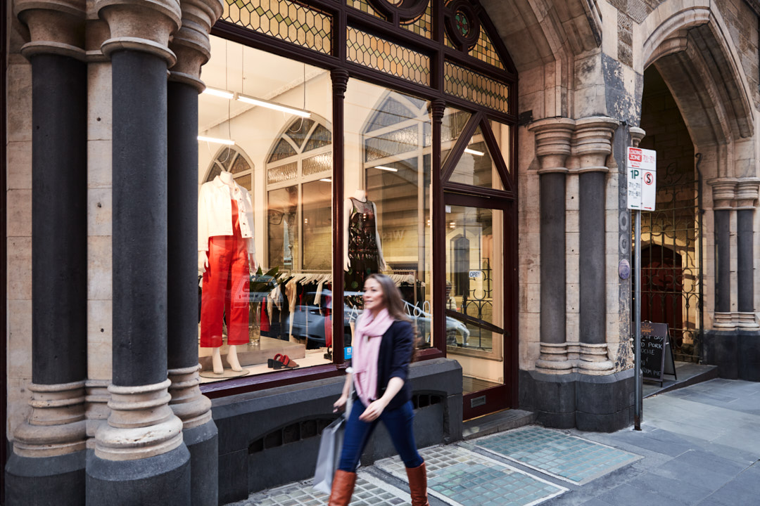 Woman walks past fashion shop window in city. Mannequins display red and white outfits. Architectural details add to urban scene.