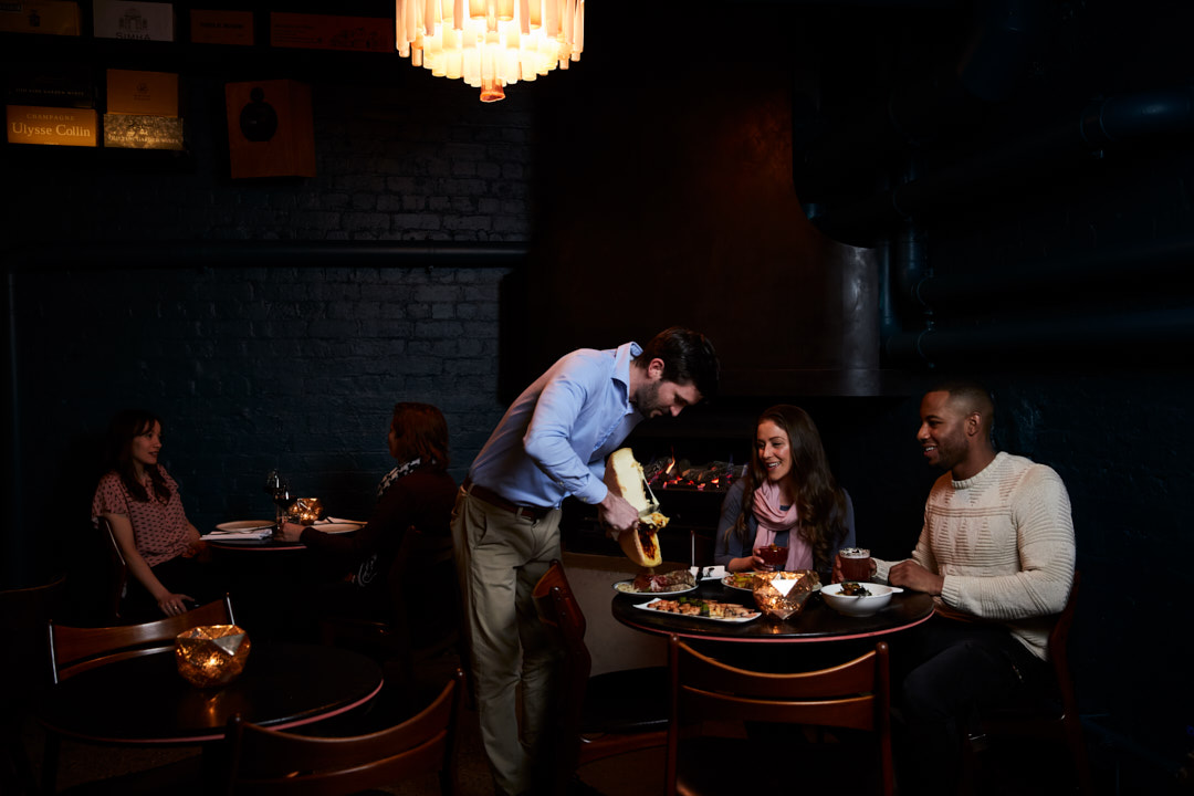In a dimly lit restaurant, a waiter serves cheese to a diverse group of seated patrons at a table. Warm ambient lighting creates an inviting atmosphere.