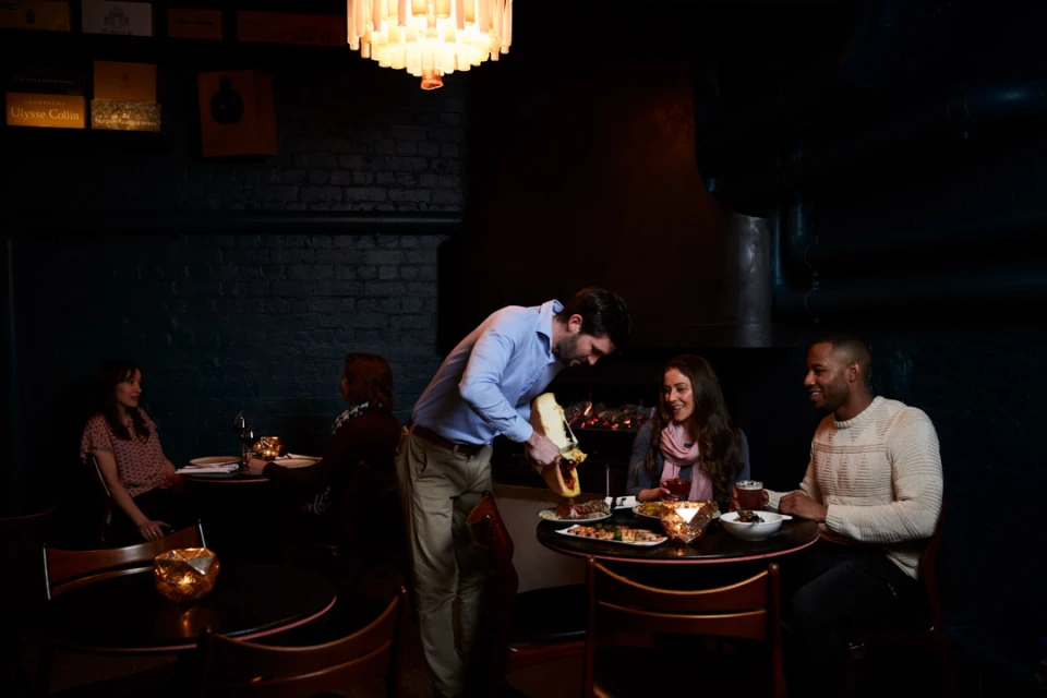 In a dimly lit restaurant, a waiter serves cheese to a diverse group of seated patrons at a table. Warm ambient lighting creates an inviting atmosphere.