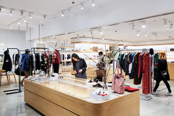 Brightly lit clothing store interior, with diverse clothing racks, tables displaying shoes and accessories, and several shoppers browsing.