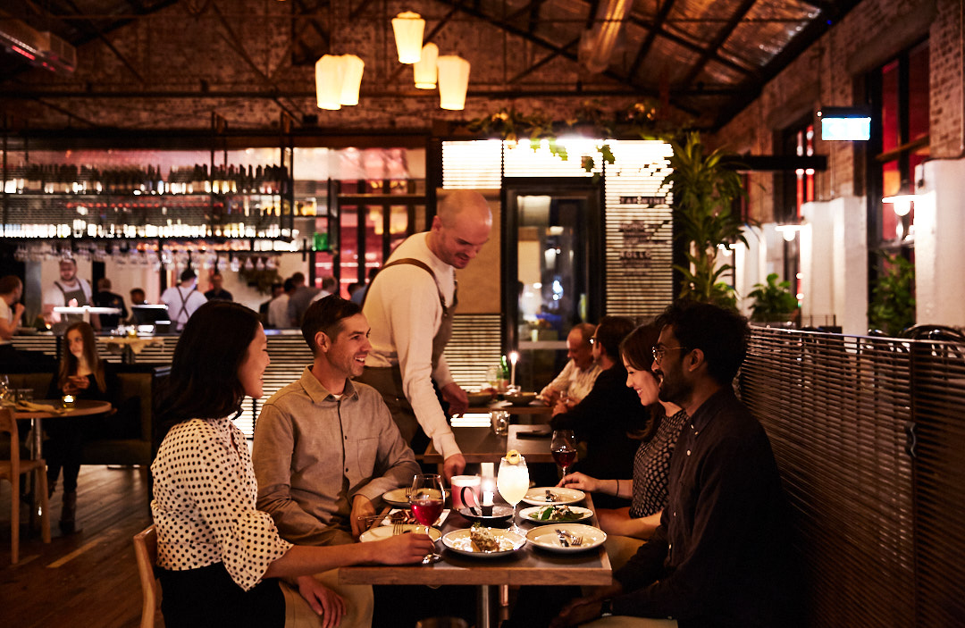 People dining in a restaurant. Customers sit at tables and a server walks between them. Interior features warm lighting and a modern design.
