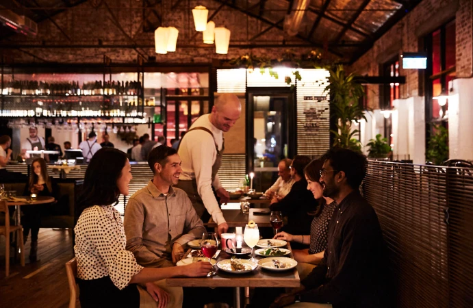 People dining in a restaurant. Customers sit at tables and a server walks between them. Interior features warm lighting and a modern design.