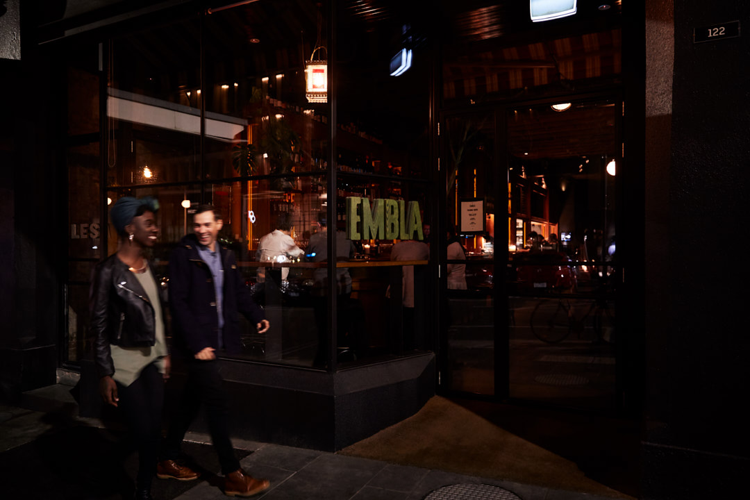 A smiling couple walks past a restaurant at night. The woman wears a leather jacket, and the man wears a coat as they walk on the sidewalk.