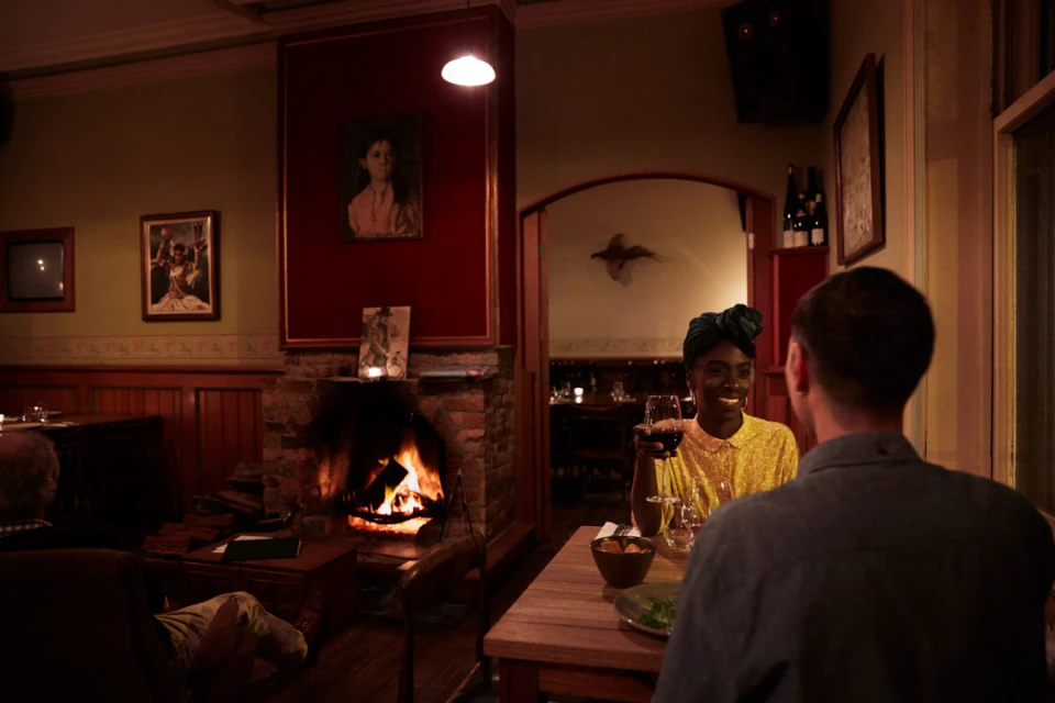 Woman in yellow dress and headscarf smiles while holding wine glass across table from man in dimly lit restaurant, fireplace in background.
