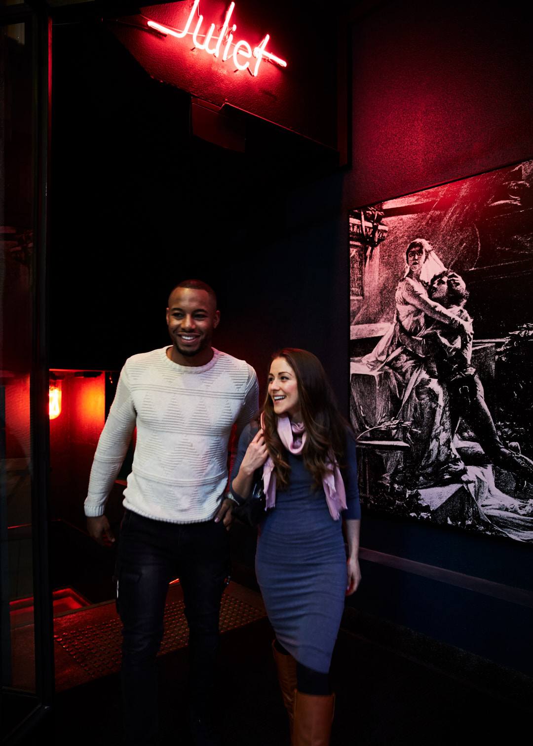 A smiling African-American man and a smiling Caucasian woman walk together in dim light, with a neon sign and artwork behind them.