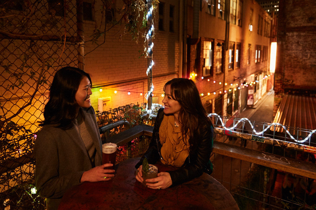 Two women at an outdoor bar at night, talking over drinks. Fairy lights and apartment buildings in the background create a warm atmosphere.