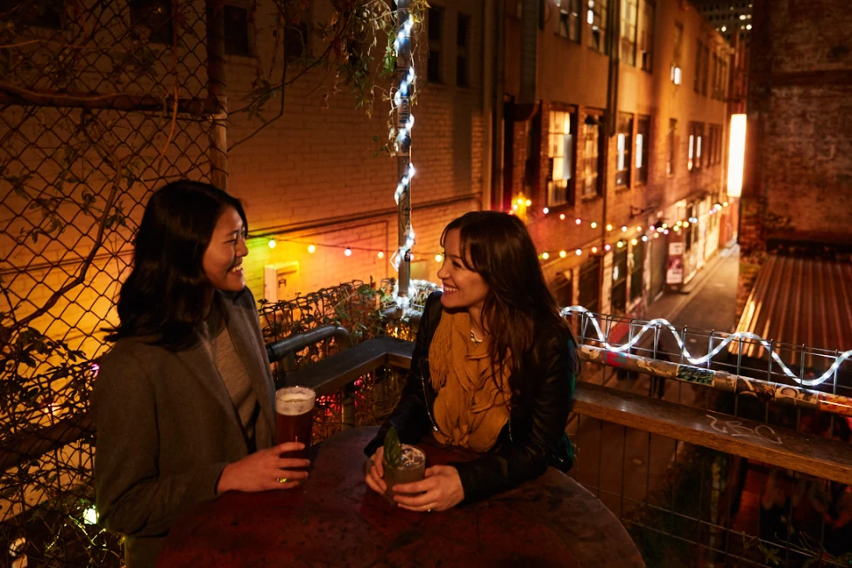 Two women at an outdoor bar at night, talking over drinks. Fairy lights and apartment buildings in the background create a warm atmosphere.