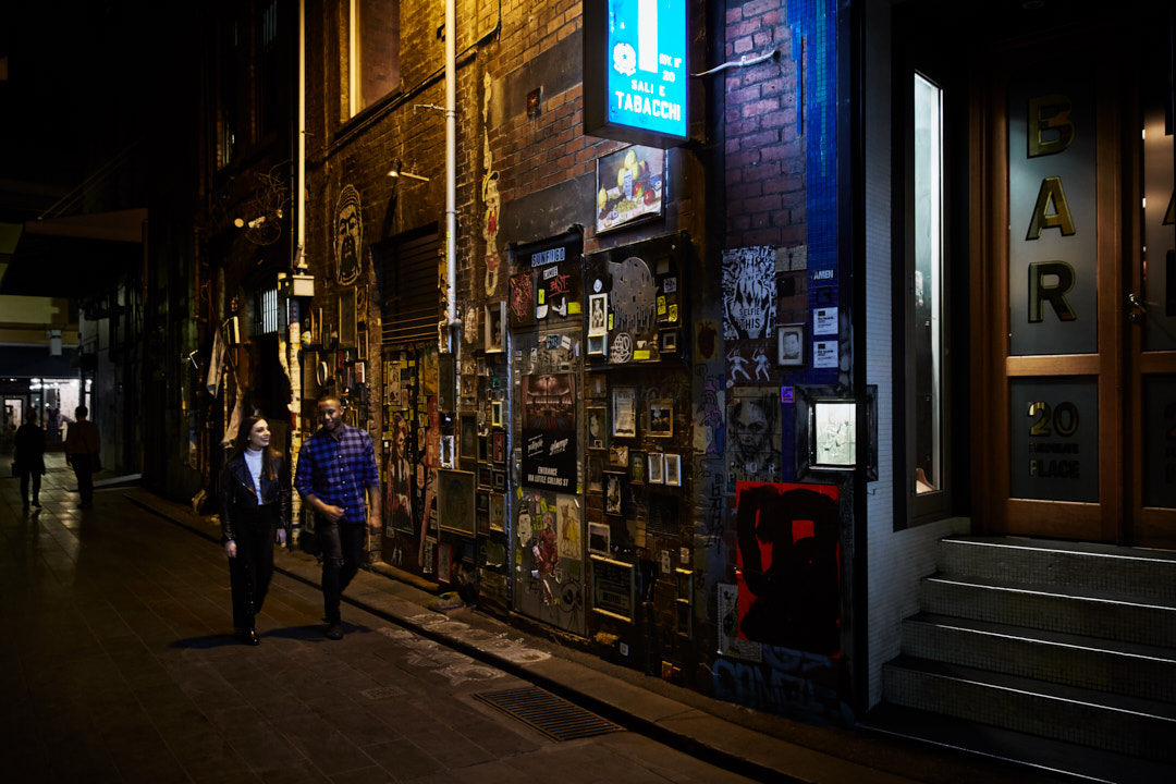 Nighttime urban scene featuring a couple walking along a narrow alleyway adorned with graffiti and posters next to a bar entrance.