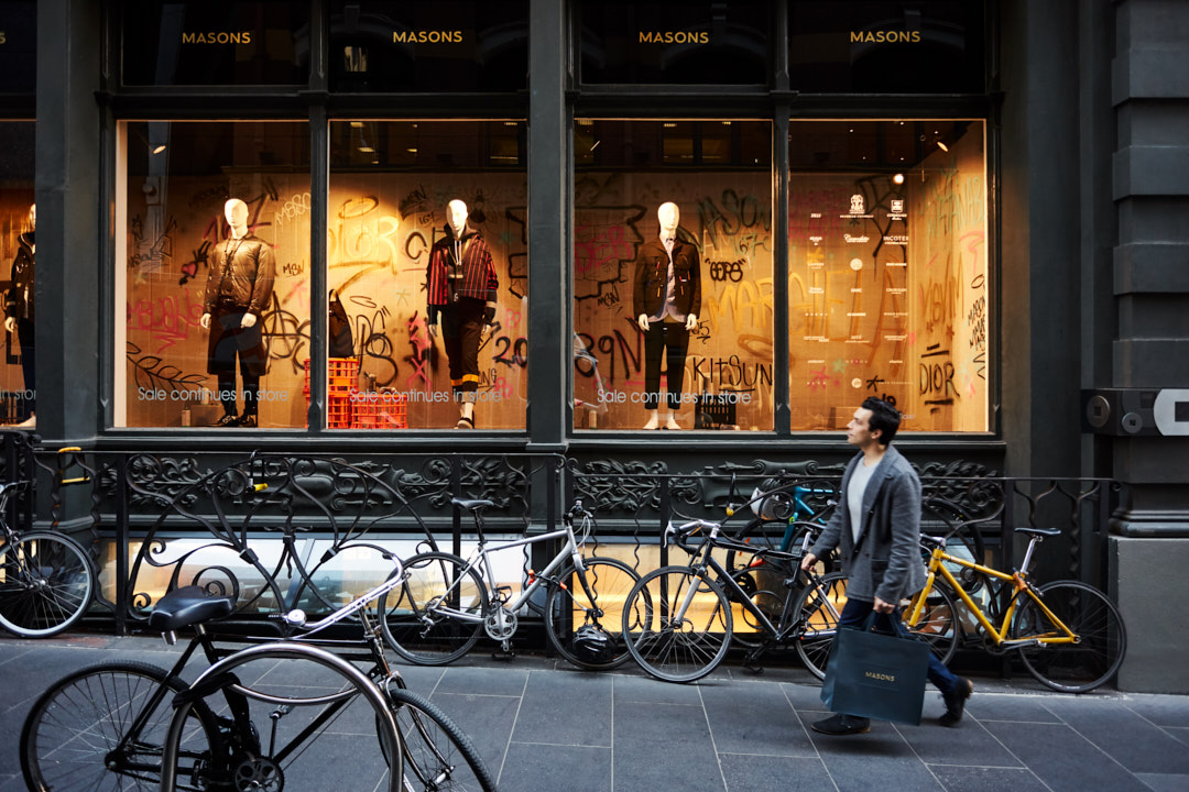 Man walks past fashion store with window display of mannequins wearing coats, jackets, and pants; bicycles parked in front of store.