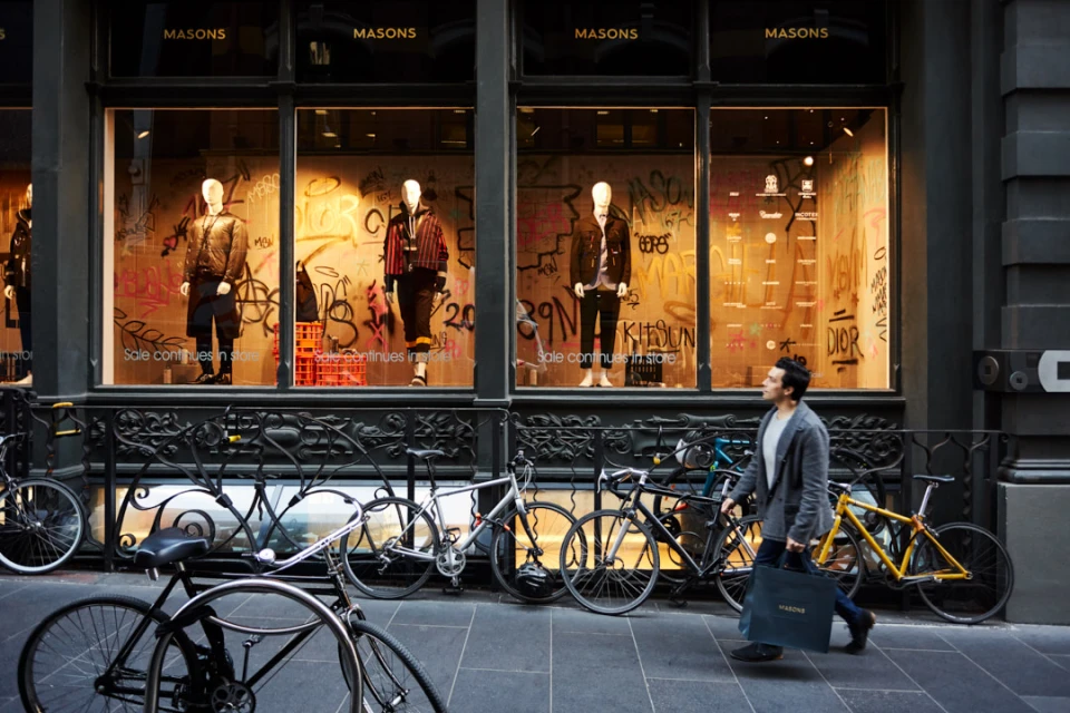 Man walks past fashion store with window display of mannequins wearing coats, jackets, and pants; bicycles parked in front of store.
