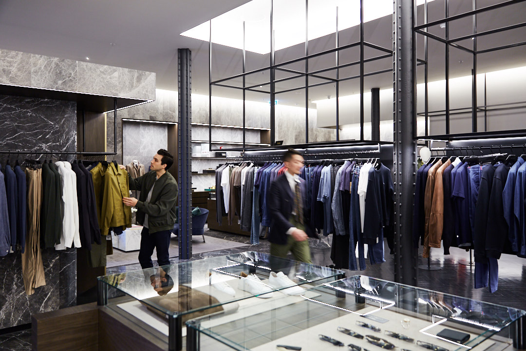 Two men browsing suits in a clothing store with racks of apparel on hangers and glass display cases in a well-lit retail environment.