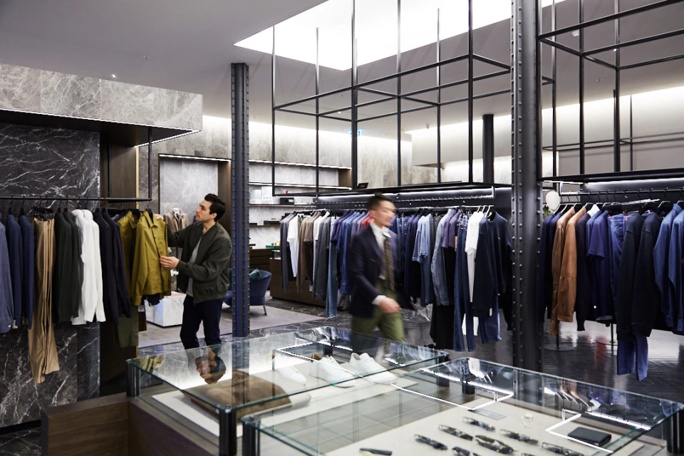 Two men browsing suits in a clothing store with racks of apparel on hangers and glass display cases in a well-lit retail environment.