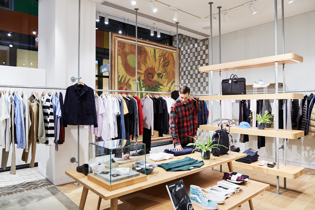 A man in a red flannel shirt browsing through a clothing store. Clothes hang on racks, while shoes and accessories displayed on tables.