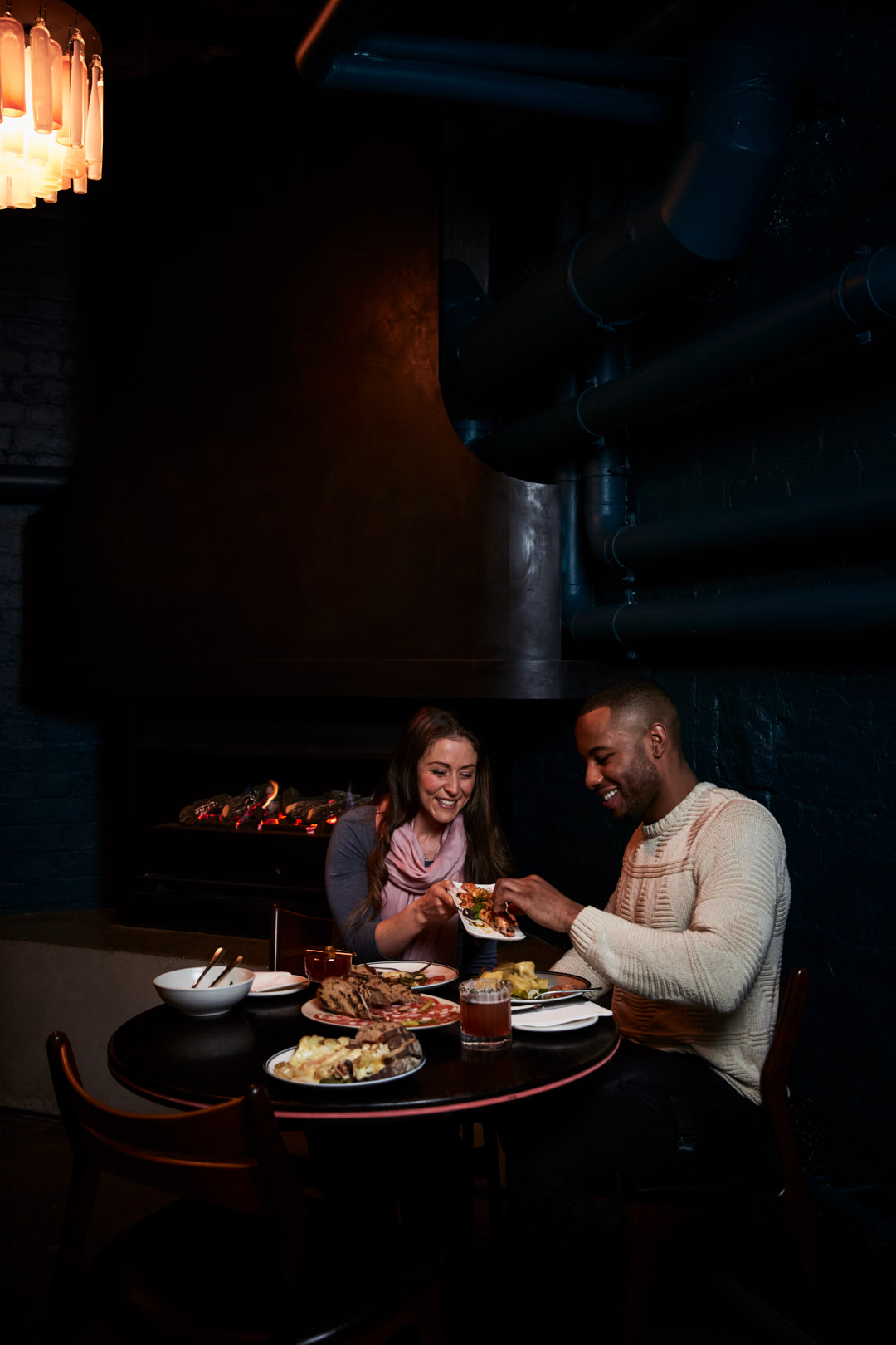 A smiling couple shares food at a candlelit table in a dark restaurant. They are sitting at a table with food plates and drinks.