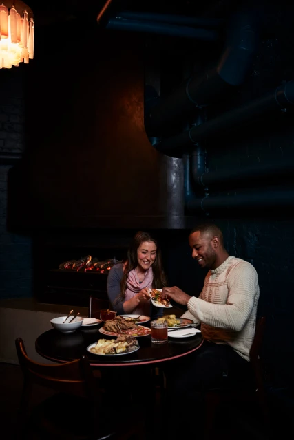 A smiling couple shares food at a candlelit table in a dark restaurant. They are sitting at a table with food plates and drinks.
