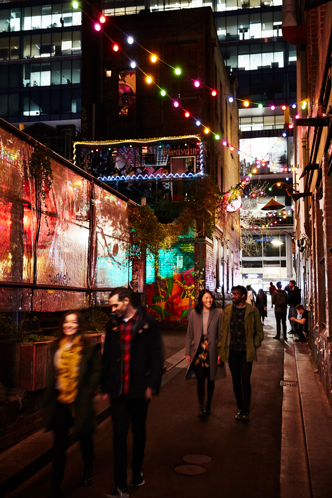 Nighttime alleyway scene with three pedestrians, brick buildings, and overhead string lights creating a warm, inviting ambiance.