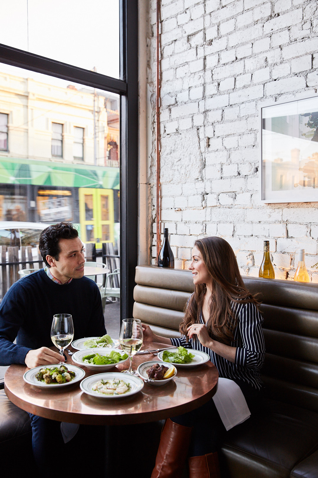 A couple enjoys a meal at a restaurant with brick walls, featuring plates of food, wine glasses, and a view through a large window.