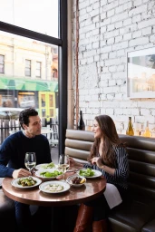 Man and woman dining at a round wooden table inside a restaurant with white exposed brick walls and large windows.