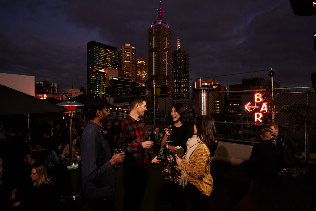 Nighttime rooftop gathering in a city with skyscrapers, featuring diverse adults in conversation and an illuminated 'Bar' sign.