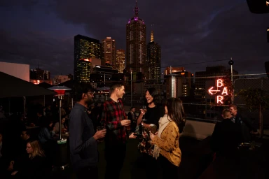 Group of friends socializing on a city rooftop bar at night with illuminated skyscrapers visible in the background.