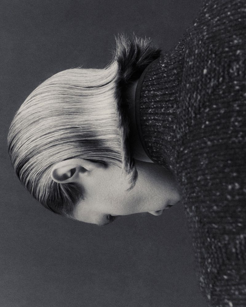 Monochrome portrait of a person with slicked-back hair and wearing a speckled sweater, presented on a gray backdrop in a studio setting.