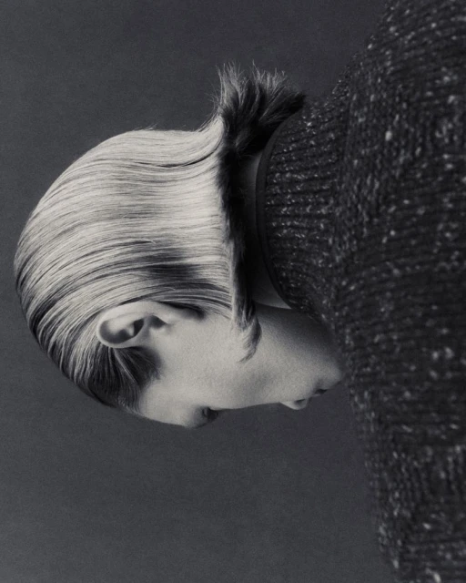 Monochrome portrait of a person with slicked-back hair and wearing a speckled sweater, presented on a gray backdrop in a studio setting.