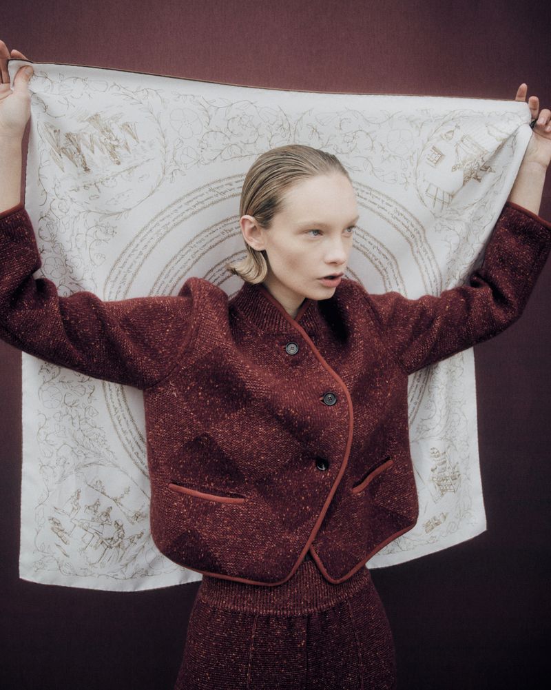 Fashionable woman in a maroon jacket holds a patterned scarf indoors, creating a stylish portrait with a muted backdrop.